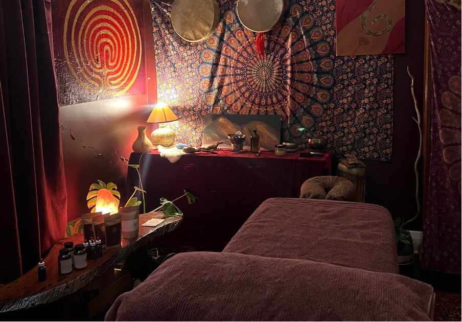 the womb massage room filled with deep reds, oranges and purples. tapestries, ceremonial drums, salt lamps, oils and herbs and feminine figures.