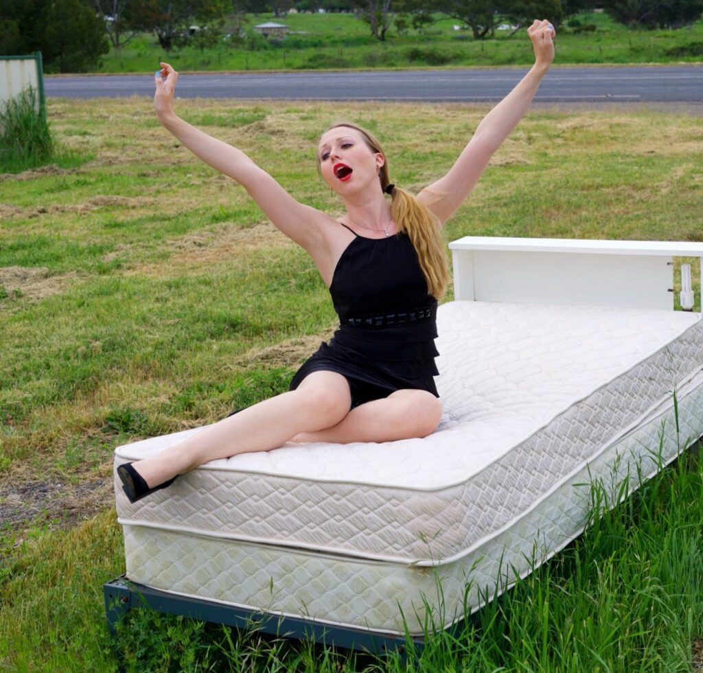 Woman with arms outstretched on mattress in grass, embracing self-love and presence now.